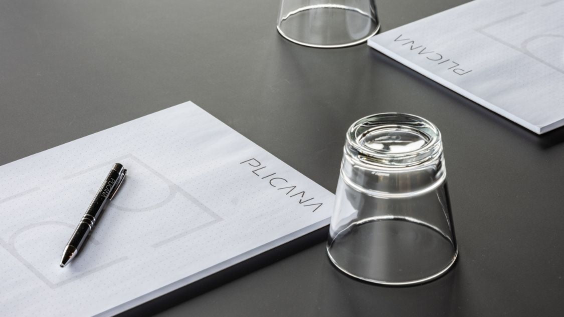 Weiße Schreibblöcke von PLICANA mit einem Kugelschreiber von PLICANA darauf und ein umgedrehtes Wasserglas