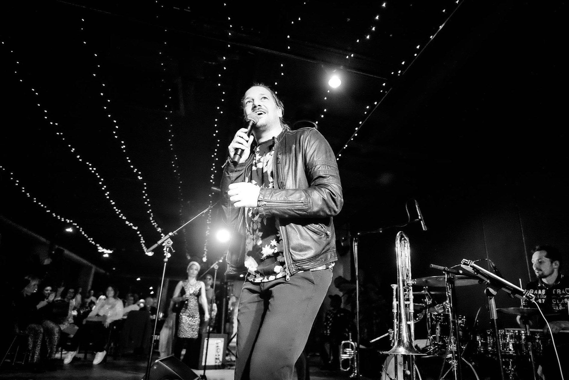 Sänger im STAGE des PLICANA in Kronau mit Band hinter ihm und Lichterketten über ihn in schwarz-weiß