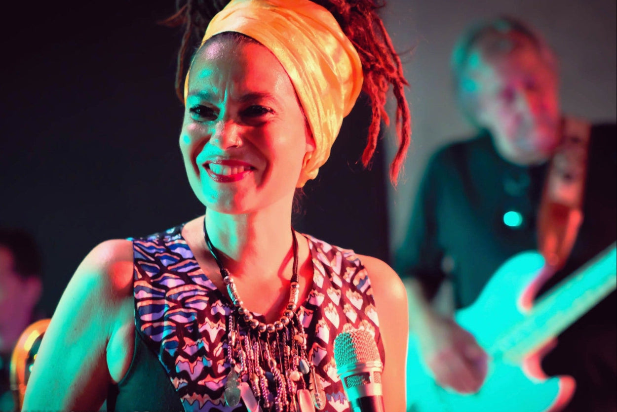 Sängerin mit Mikrofon, Blau-schwarzem Oberteil und Perlkette lächelt. Im Hintergrund ist ein Gitarrist