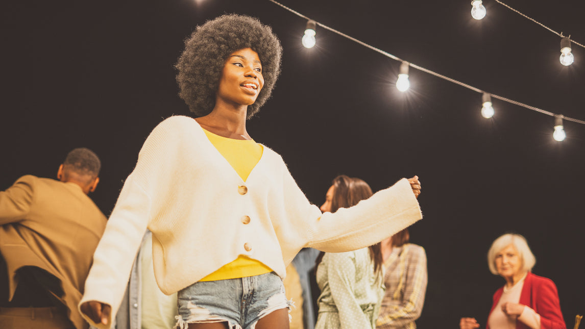 Frau mit Afro, gelbem Oberteil und weißem Cardigan tanzt Abends auf dem ROOF im PLICANA