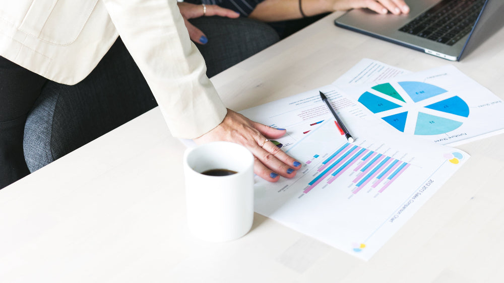 Auf weißem Tisch liegen Blätter mit Diagrammen, einer Kaffeetasse und einem Laptop. Eine Frauenhand lehnt sich an den Tisch an