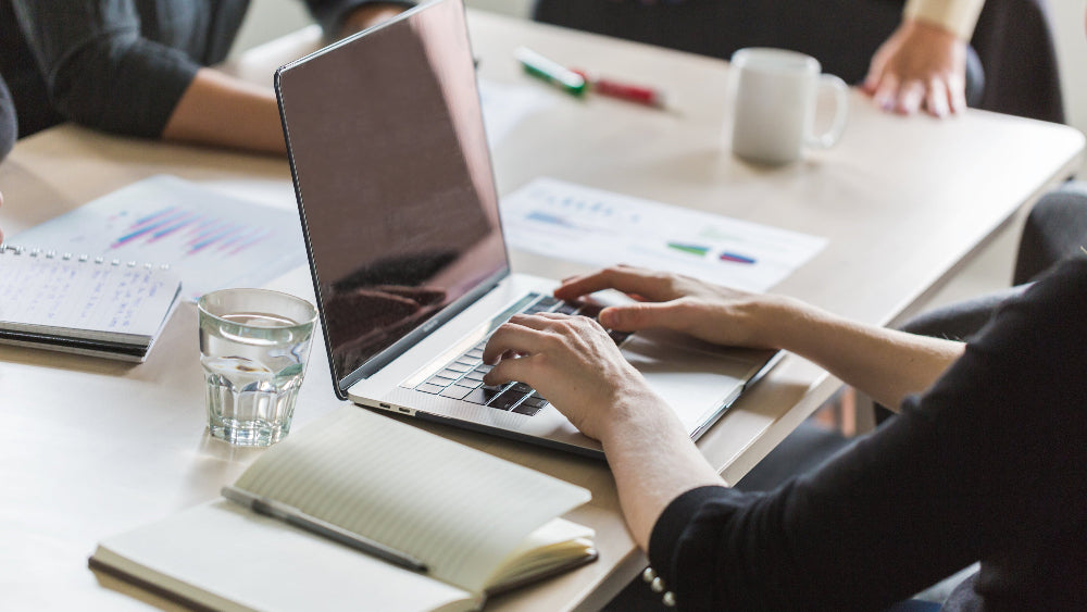 Auf weißem Tisch liegen Notizblöcke, ein Laptop und Gläser. Menschen tippen auf Laptop und schreiben in Blöcke