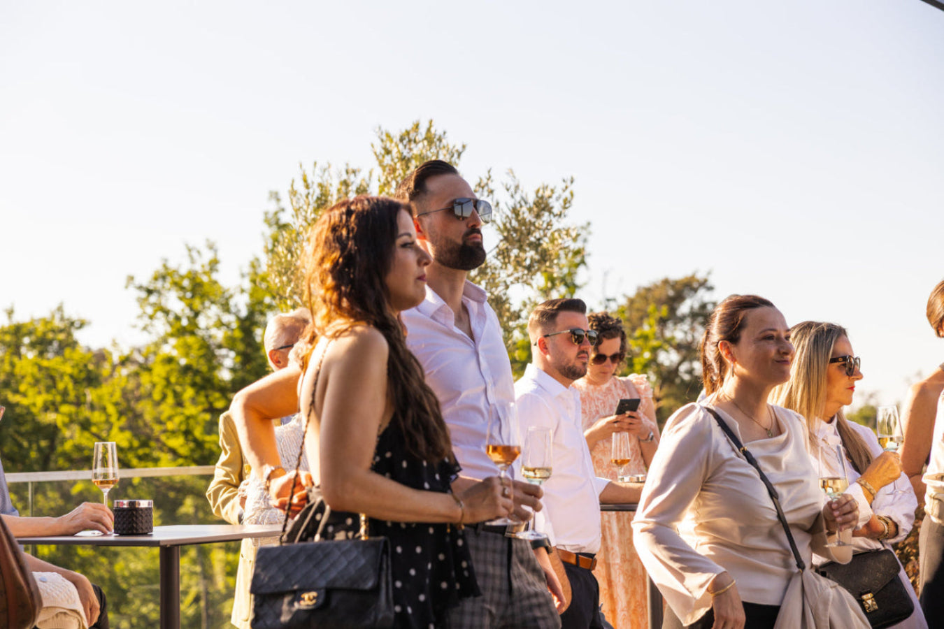Menschen auf Feier stehen um Stehtische mit Sektgläsern in der Hand schauen nach rechts. Vier Personen haben eine Sonnenbrille an